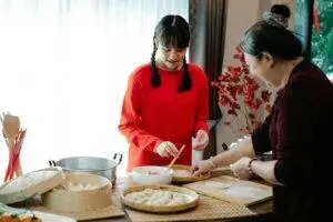 Smiling ethnic grandma with female teen preparing Chinese dumplings while talking at table with traditional steamers in house