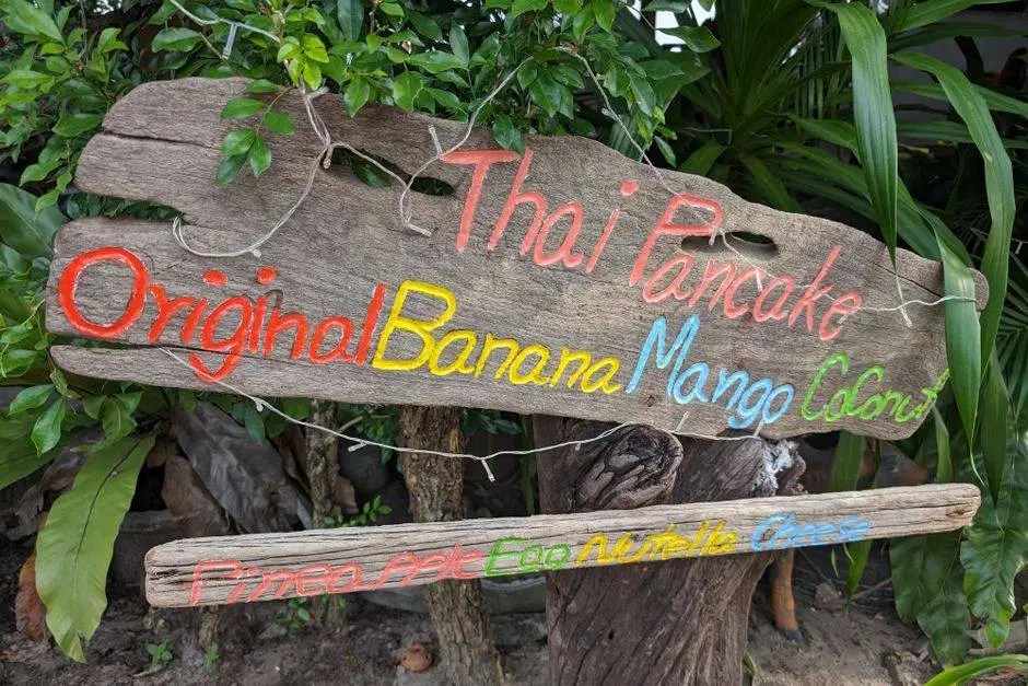 Colorful wooden sign showcasing Thai pancakes with tropical fruit flavors amidst lush greenery.