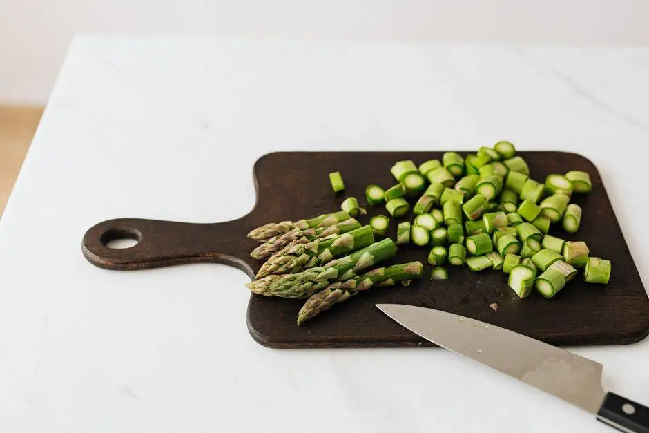 From above of chopped green asparagus near knife on wooden cutting board placed on white table